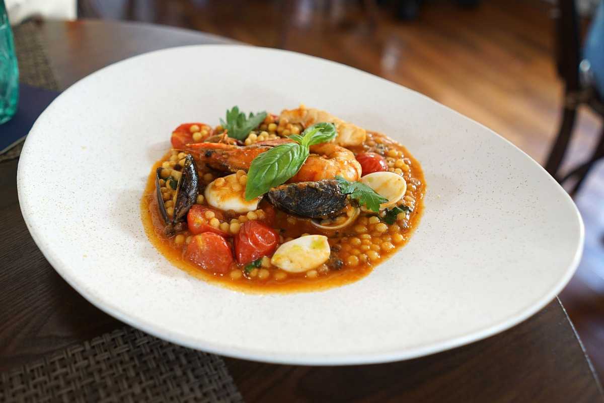 Scorcio di una tavolata all’aperto con piatti tipici di pesce, pane carasau e vino bianco, in una terrazza con vista mare a San Teodoro al tramonto