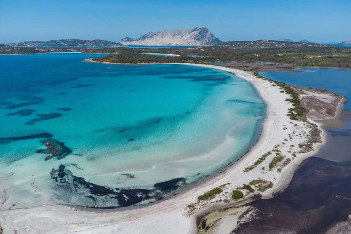 Bambini che giocano con secchiello e paletta sulla spiaggia bianca di Cala Brandinchi, alle spalle pini e mare trasparente. Atmosfera familiare e allegra.